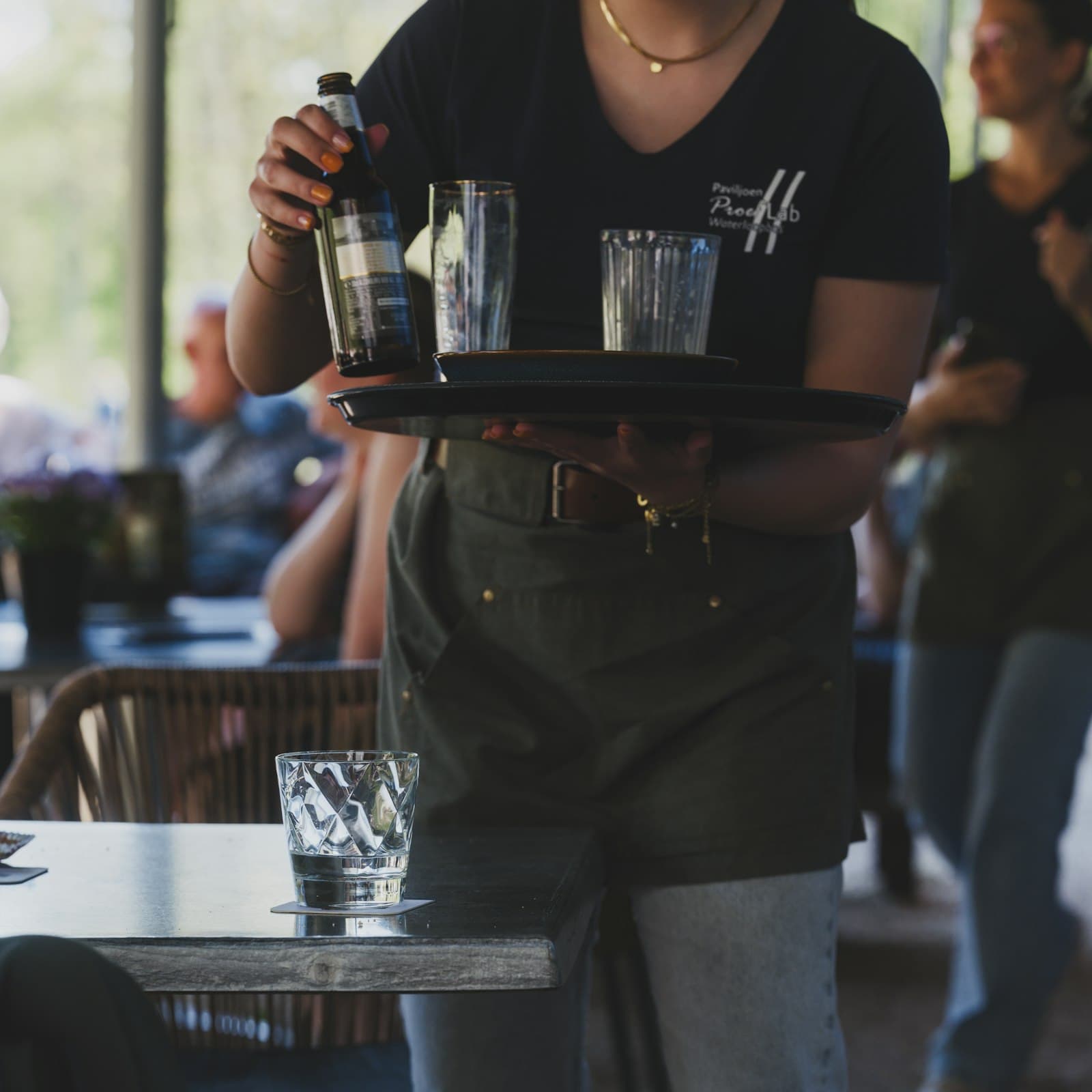 A server carrying a tray of glasses and a water bottle through a busy restaurant